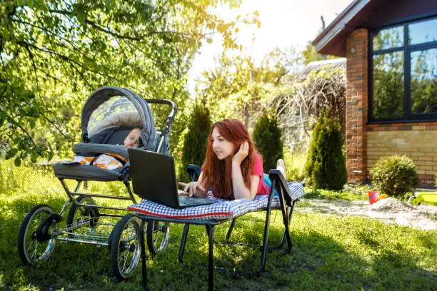 Photo of Young woman with her newborn baby lying on the soft armchair with laptop. Summer day. Mother with child outdoors. Motherhood.