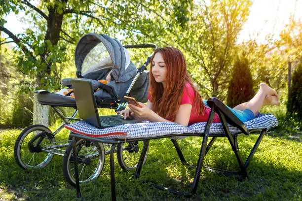 Photo of Young woman with her newborn baby lying on the soft armchair with laptop and looking at the phone. Summer day. Mother with child outdoors. Motherhood.