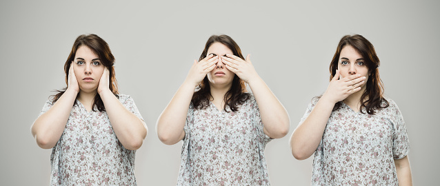 Montage of a young caucasian woman covering her eye, ears and mouth with hands. Horizontal portrait of real woman gesturing hear no evil, see no evil and speak no evil against gray background. Studio photography from a DSLR camera. Sharp focus on eyes.