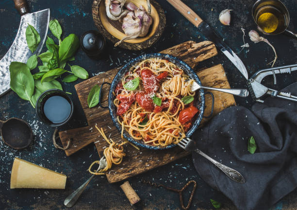 Spaghetti with tomato and basil and ingredients for making pasta Italian style pasta dinner. Spaghetti with tomato and basil in plate on wooden board and ingredients for cooking pasta over dark blue plywood background, top view pasta photos stock pictures, royalty-free photos & images