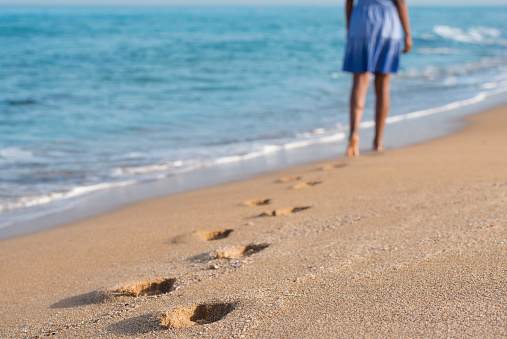 Close up image on woman's footprints on yellow sea sand. Unrecognized woman walking down beach, wearing blue dress.