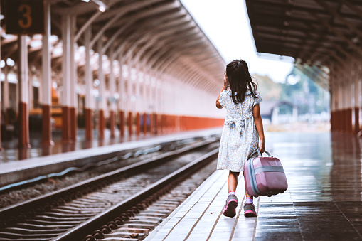 Back view of cute asian little girl holding suitcase and walking on a railway station in vintage retro style