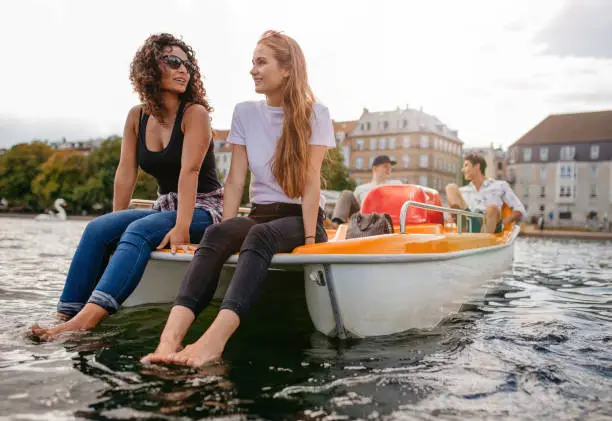 Shot of two women sitting in front pedal boat with feet in water and man in background. Teenage friends enjoying boating in the lake.