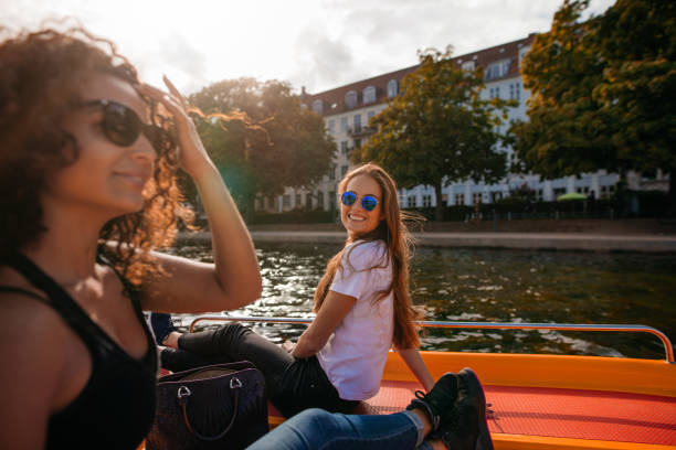Shot of smiling young woman sitting on deck of boat with female friend in front. Two young women on boat in lake.