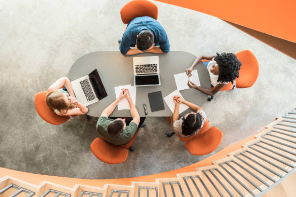View from above towards five business people around meeting table Young women and men sitting on orange chairs in discussion with laptops and notes on table directly above stock pictures, royalty-free photos & images