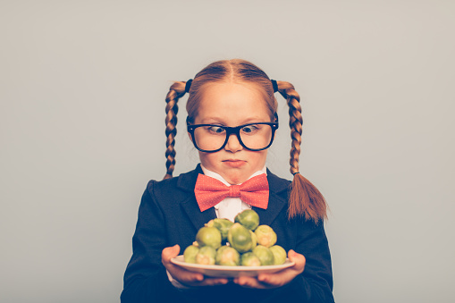 A young girl nerd wearing eyeglasses, bow tie and cardigan holds a plate of brussels sprout. She is alarmed at the thought of having to eat her vegetables. Holding them and looking at them will not help.