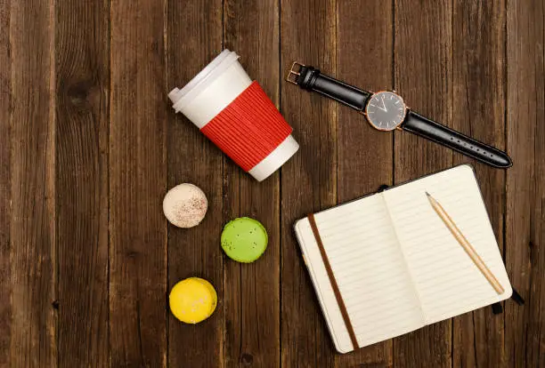 Open notebook with a pencil, macaroons, paper-cups and a watches on a wooden background. Top view