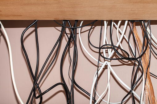 Untidy cables hanging behind a computer desk. Various colors and types of cables hanging loose and tangled without cable management.