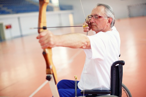 Older paraplegic man in wheelchair aiming with bow and arrow on archery training