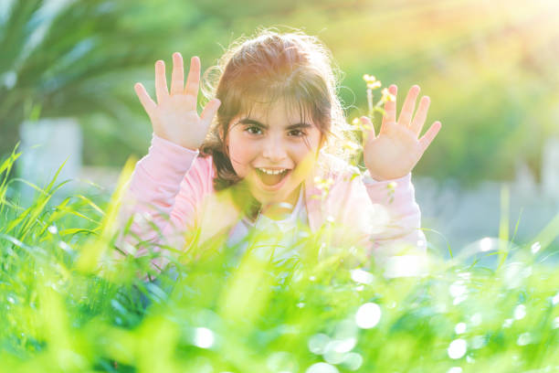 Pretty child playing outdoors, cute little girl raised up hands and laughing, having fun on the fresh green grass field in bright sun light, enjoying nice spring day
