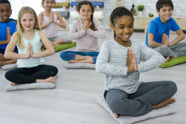 We Love Yoga Class Cute young elementary age girl of african descent wearing a grey shirt sitting on a cushion smiling and holding prayer pose with a multi-ethnic group of kids during a yoga class in a bright white studio. mindfulness kids stock pictures, royalty-free photos & images