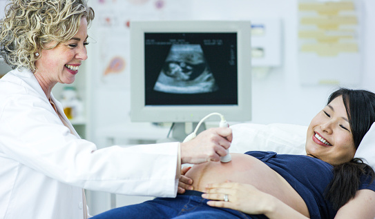 Smiling caucasian female doctor using an ultrasound doppler on the stomach of a beautiful asian pregnant patient during a routine prenatal checkup at the doctor's office in a medical clinic.