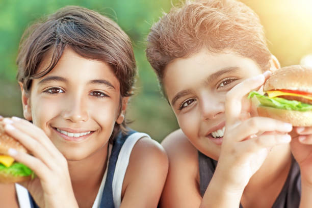 Portrait of a two happy boys eating hamburgers outdoors, cheerful teens enjoying unhealthy but delicious food, picnic on sunny day