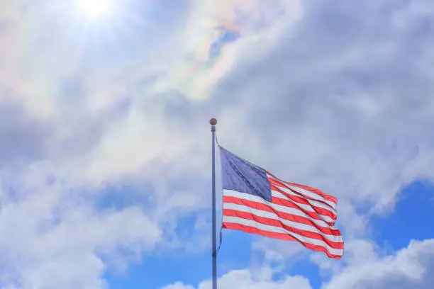 American Flag blowing in wind against bright sun, partly cloudy sky