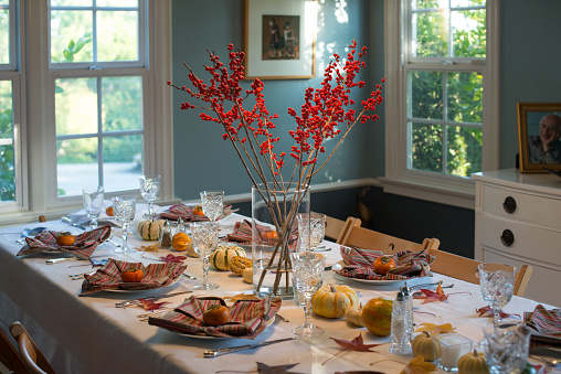 Dining room table completely decorated for Thanksgiving with colors of orange, white, brown, yellow, red with items such as pumpkins, branches with berries, colorful leaves on  top of an all white table cloth. Table is fully set with table settings, cutlery, wine glasses, salt and pepper shakers and napkins.