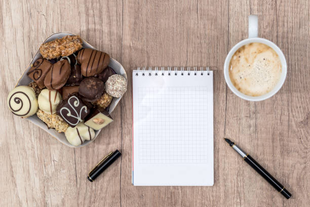 Close up of a bowl of biscuits and several cups on a table, next to female hands writing something in a notebook, open laptop in blurred background.
