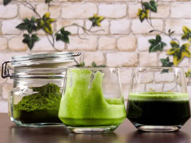 Two glasses of green juice, one full and one empty, plus one jar of green juice powder, set over a wenge board, with a background of brick wall and ivy leafs.