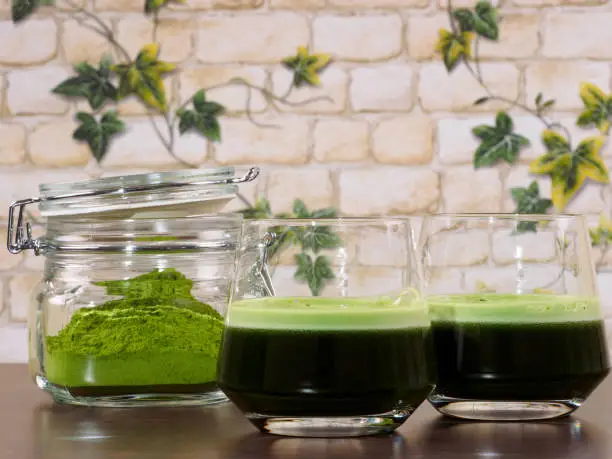 Two glasses of green juice and a jar of green juice powder, set over a wenge board, with a background of brick wall and ivy leafs