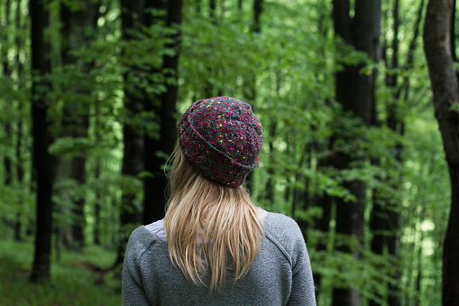 horizontal rear view portrait of Caucasian young woman with long blonde hair and colorful wool hat standing in a green forest