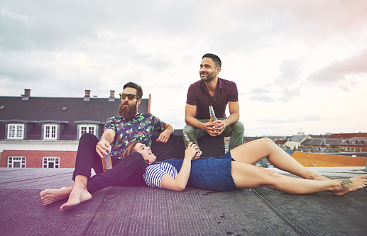 Singing young woman laying in lap of bearded man near friend on roof in European city drinking bottles of beer