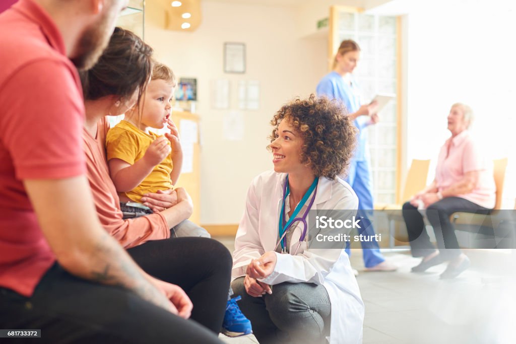 Toddler talking to female doctor A young family sits talking to the doctor. The toddler is sat on his mother’s knee as the doctor kneels down to talk to him Doctor Stock Photo