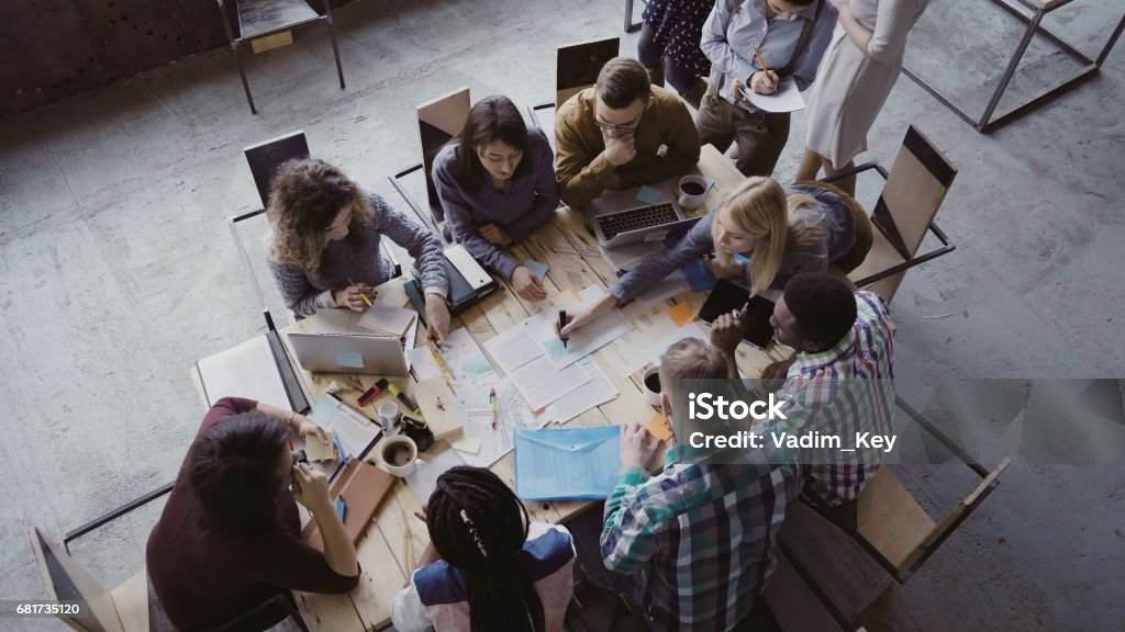 Top view of mixed race business team sitting at the table at loft office and working. Woman manager brings the document Top view of creative business team sitting at the table at loft office and working. Woman manager brings the document to mixed race group of people. Colleagues discussing the project. Meeting Stock Photo