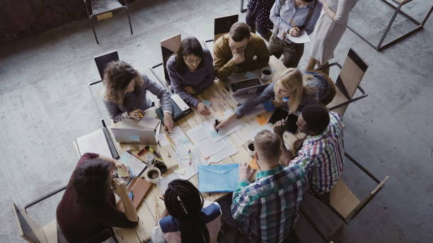 Top view of mixed race business team sitting at the table at loft office and working. Woman manager brings the document Top view of creative business team sitting at the table at loft office and working. Woman manager brings the document to mixed race group of people. Colleagues discussing the project. belarus stock pictures, royalty-free photos & images