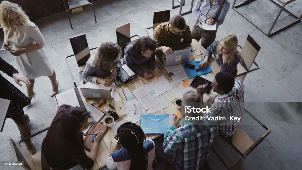 Top view of mixed race business team sitting at the table at loft office and working. Woman manager brings the document Top view of creative business team sitting at the table at loft office and working. Woman manager brings the document to mixed race group of people. Colleagues discussing the project. Casual Clothing Stock Photo