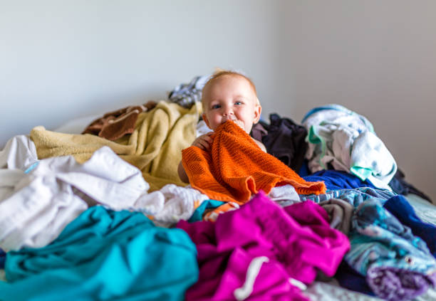 Cute Adorable Baby Sits in a Pile of Laundry on the Bed Young caucasian boy disrupts house chores by climbing onto bed on top of laundry. baby clothing stock pictures, royalty-free photos & images