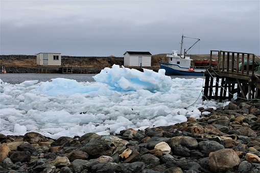 Pack Ice in Ferryland, Newfoundland and Labrador, Canada, April 22, 2017. The ice is right to the beach on one side of the inlet but a boat sits in open water on the far side. A rock beach, a pier, and some outbuildings are also shown.