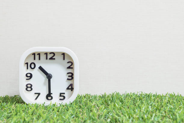 istock Closeup white clock for decorate show a half past ten, or 10:30 a.m. on green artificial grass floor and cream wallpaper textured background with copy space 682060718