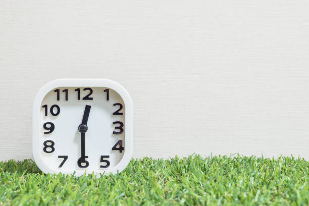 istock Closeup white clock for decorate show a half past twelve , or 12:30 a.m. on green artificial grass floor and cream wallpaper textured background with copy space 682060904