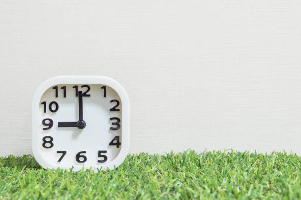 istock Closeup white clock for decorate in 9 o'clock on green artificial grass floor and cream wallpaper textured background with copy space 682061100