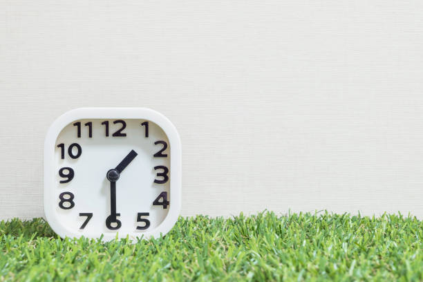 istock Closeup white clock for decorate show a half past one or 1:30 p.m. on green artificial grass floor and cream wallpaper textured background with copy space 682061238