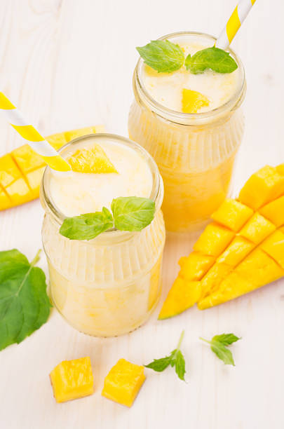 istock Freshly blended yellow mango fruit smoothie in glass jars with straw, mint leaves, mango slices, close up. Soft white wooden board background. 682089392