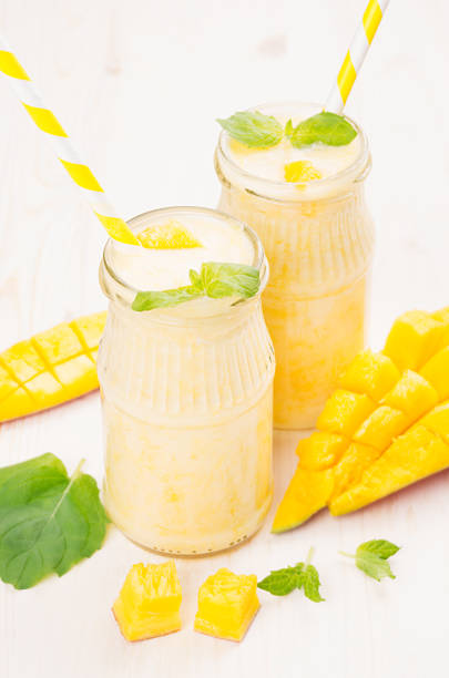 istock Yellow mango fruit smoothie in glass jars with straw, mint leaves, mango slices, close up. Soft white wooden board background. 682089402