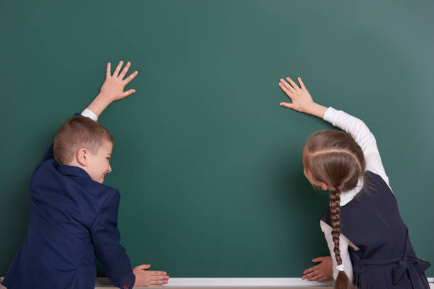 istock elementary school boy and girl put hands on chalkboard background and show blank space, dressed in classic black suit, group pupil, education concept 682126122