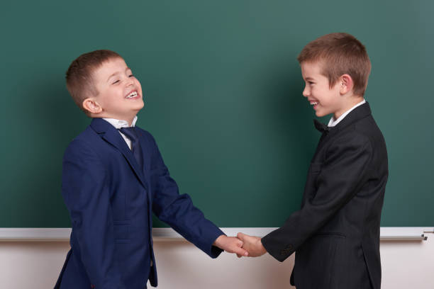 istock two friends handshake, elementary school boy near blank chalkboard background, dressed in classic black suit, group pupil, education concept 682126238