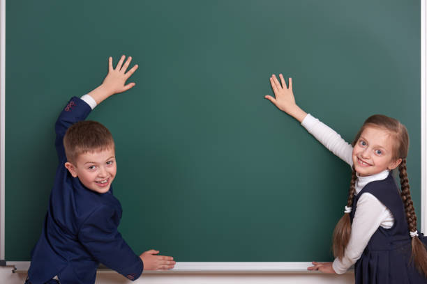 istock elementary school boy and girl put hands on chalkboard background and show blank space, dressed in classic black suit, group pupil, education concept 682126546