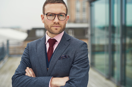 Single serious handsome bearded young businessman in eyeglasses, red necktie and folded arms outside on balcony at office