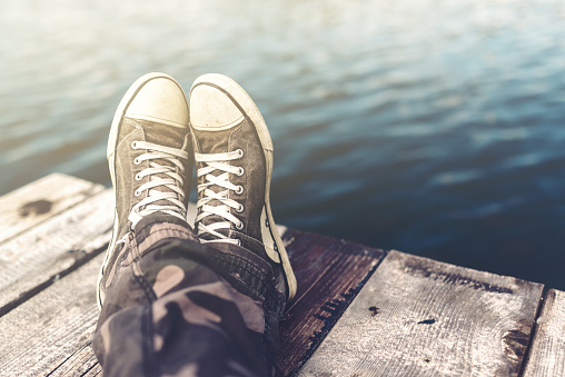 Man with crossed legs relaxing on wooden riverbank pier, casual young guy wearing sneakers sitting by the river in summer afternoon, selective focus