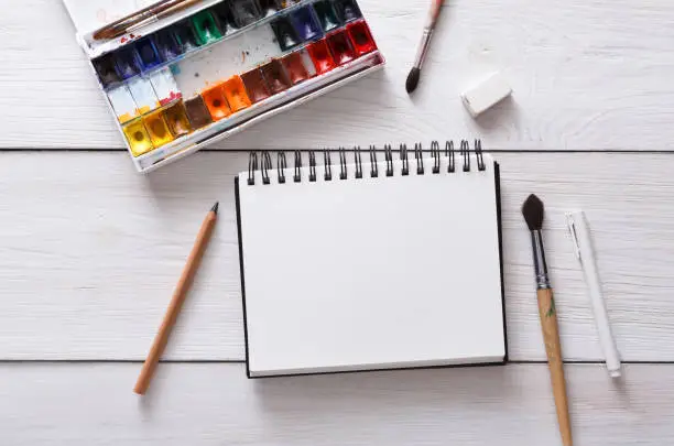 Drawing tools, stationary supplies, workplace of artist. Watercolor paints and blank paper on white wooden desk, top view, flat lay, copy space