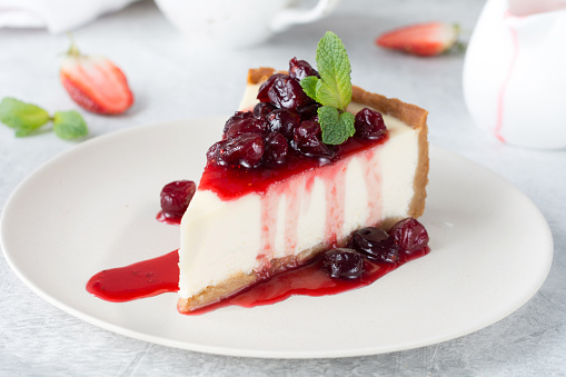 Slice of plain cheesecake with cranberry sauce on white plate decorated with mint leaf. Closeup view