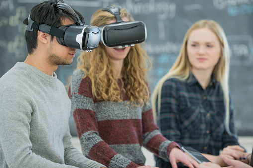 A multi-ethnic group of high school students are indoors at school. They are in a technology classroom. They are wearing casual clothing. A Caucasian girl and Ethnic boy are using virtual reality headsets. They are smiling and having fun.