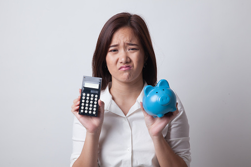 Unhappy Asian woman with calculator and piggy bank on white background