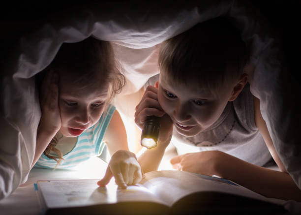 istock Sister and brother are reading a book under a blanket with flashlight. 682366612