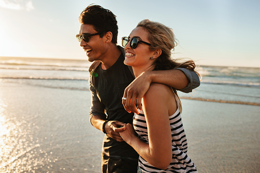 Outdoor shot of smiling young couple walking on beach. Young man and woman strolling together on seashore on a summer day.