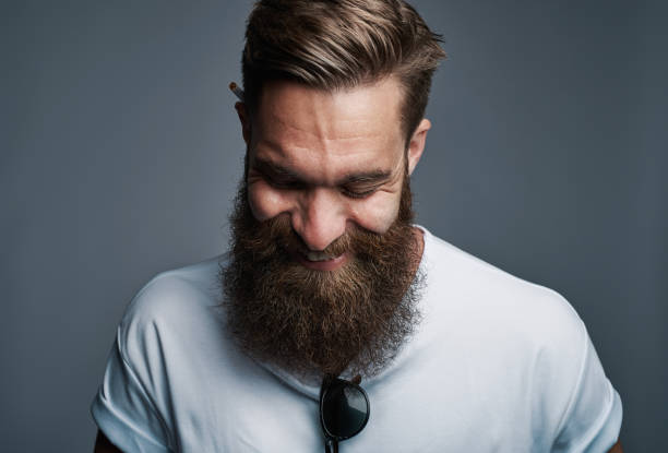 Giggling young man with large fuzzy beard Single giggling attractive young European man with large furry beard with eyeglasses placed in shirt over gray background beard stock pictures, royalty-free photos & images