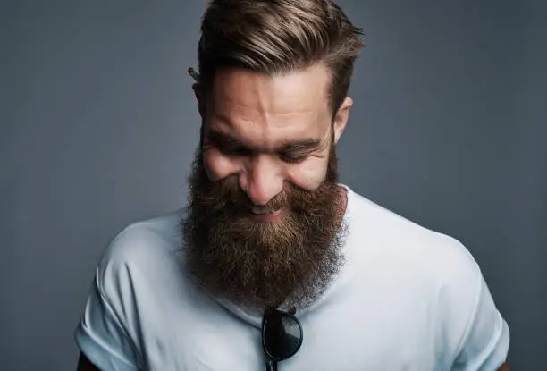 Single giggling attractive young European man with large furry beard with eyeglasses placed in shirt over gray background