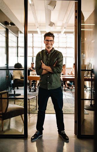 Full length portrait of smiling young man standing in doorway of office with his arms crossed. Creative male executive at startup with people working in background.